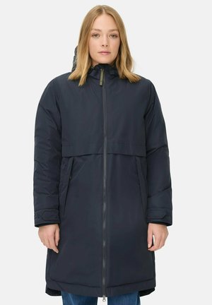 Vrouw in een lange, donkerblauwe winterjas met een rits aan de voorkant en een capuchon, staande tegen een effen achtergrond.