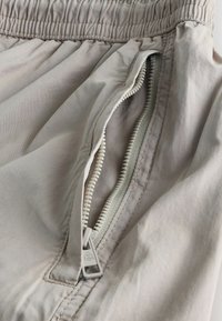 Jasnobeżowe spodnie z tkaniny z elastycznym paskiem w talii, wyposażone w częściowo otwartą kieszeń na suwak z widocznymi detalami przeszyć.