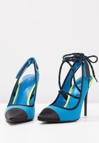 Paire de chaussures à talons hauts bleues avec embouts noirs, bordure noire et lacets noirs et bleus noués autour de la cheville sur une surface blanche.