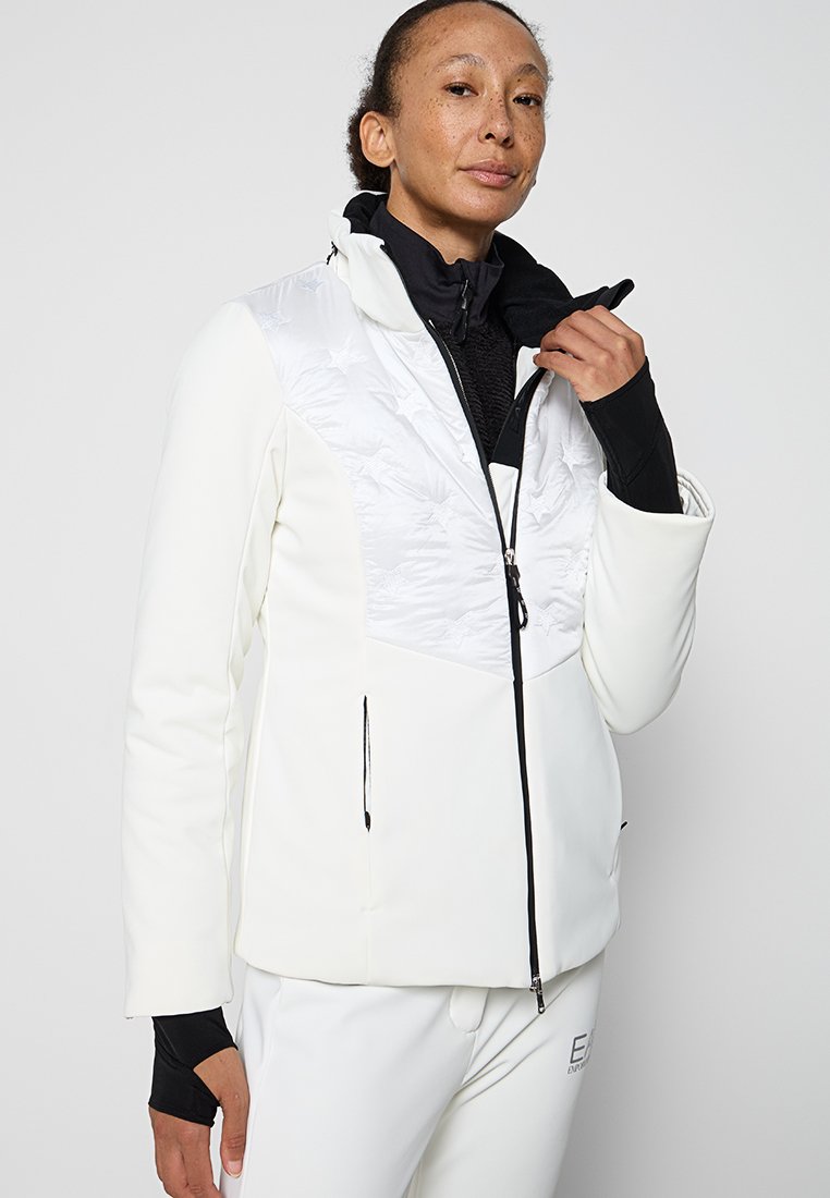 Giacca bianca con parte anteriore trapuntata, dettagli neri, colletto alto e tasche con zip. Presenta una texture liscia e un design aderente.