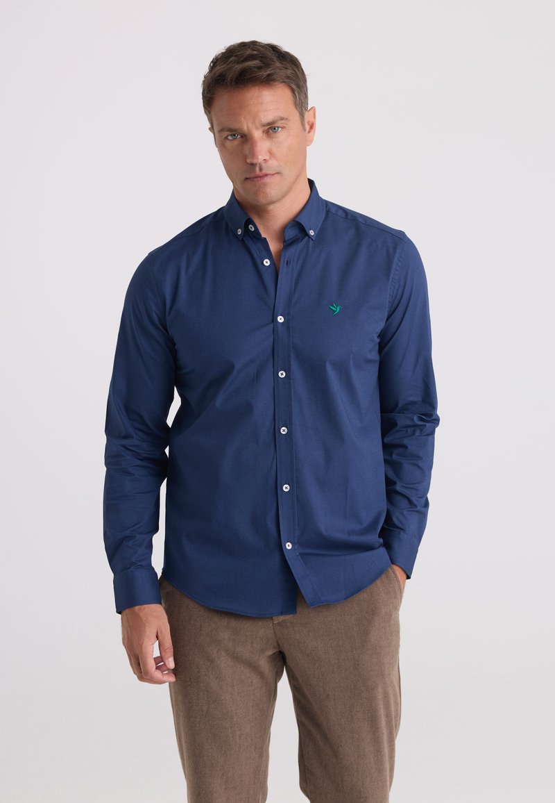 Homme portant une chemise bleu foncé boutonnée avec un petit logo vert et un pantalon marron clair, debout avec une main dans la poche.
