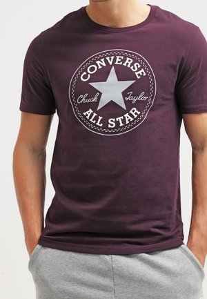 T-shirt en coton marron avec un motif d'étoile argentée accompagné du texte "Converse Chuck Taylor All Star" et une bordure circulaire. Manches courtes, col rond.
