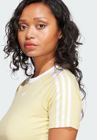 Light yellow fitted t-shirt with white three-stripe accents on sleeves and small white logo on the chest. Smooth cotton material.