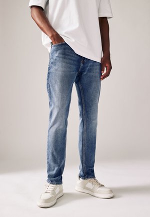 Personne portant un jean en denim bleu clair, des baskets blanches et une chemise blanche à manches courtes avec une main dans la poche.
