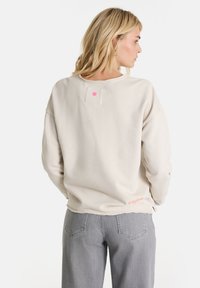 Blonde Frau in einem hellbeigen Sweatshirt und grauen Jeans, mit dem Rücken zur Kamera und den Händen in den Taschen, vor einem weißen Hintergrund.