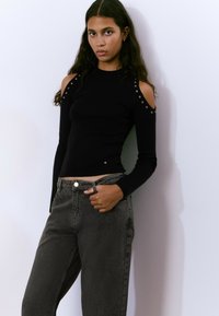 Mujer joven con cabello largo y oscuro, vistiendo un suéter negro de hombros descubiertos y jeans gris oscuro, de pie contra una pared blanca.