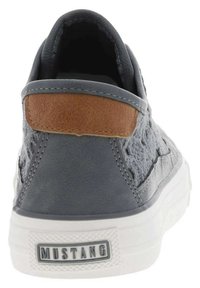 Sneaker grigio in pelle senza lacci con un accento marrone in camoscio sul tallone, suola in gomma e pannello laterale testurizzato. Presenta un logo "MUSTANG" sul retro.