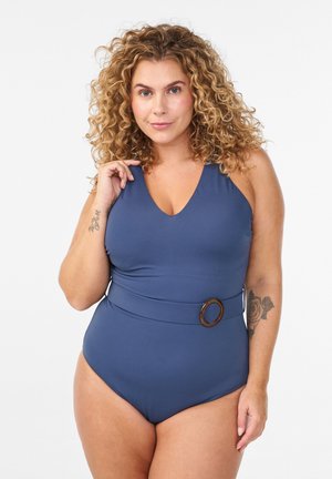 Lockige Frau trägt einen blauen, mit Gürtel versehenen Einteiler-Badeanzug, zeigt Tätowierungen am linken Arm und steht vor einem einfarbigen weißen Hintergrund.