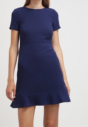 Mujer vistiendo un vestido azul marino de manga corta, por encima de la rodilla, con un dobladillo ligeramente con volantes, sobre un fondo liso.