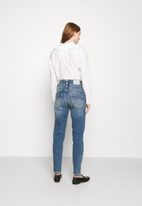 Jeans azuis desbotados com uma cintura alta, pernas ajustadas e bolsos traseiros distintos, combinados com uma camisa branca de botão. Sapatos pretos visíveis.