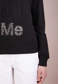 Svart sweatshirt med upphöjd text, prydd med glittriga rhinstenar som stavar "Me." Ribbstickade muddar och nederkant; parat med vita byxor.