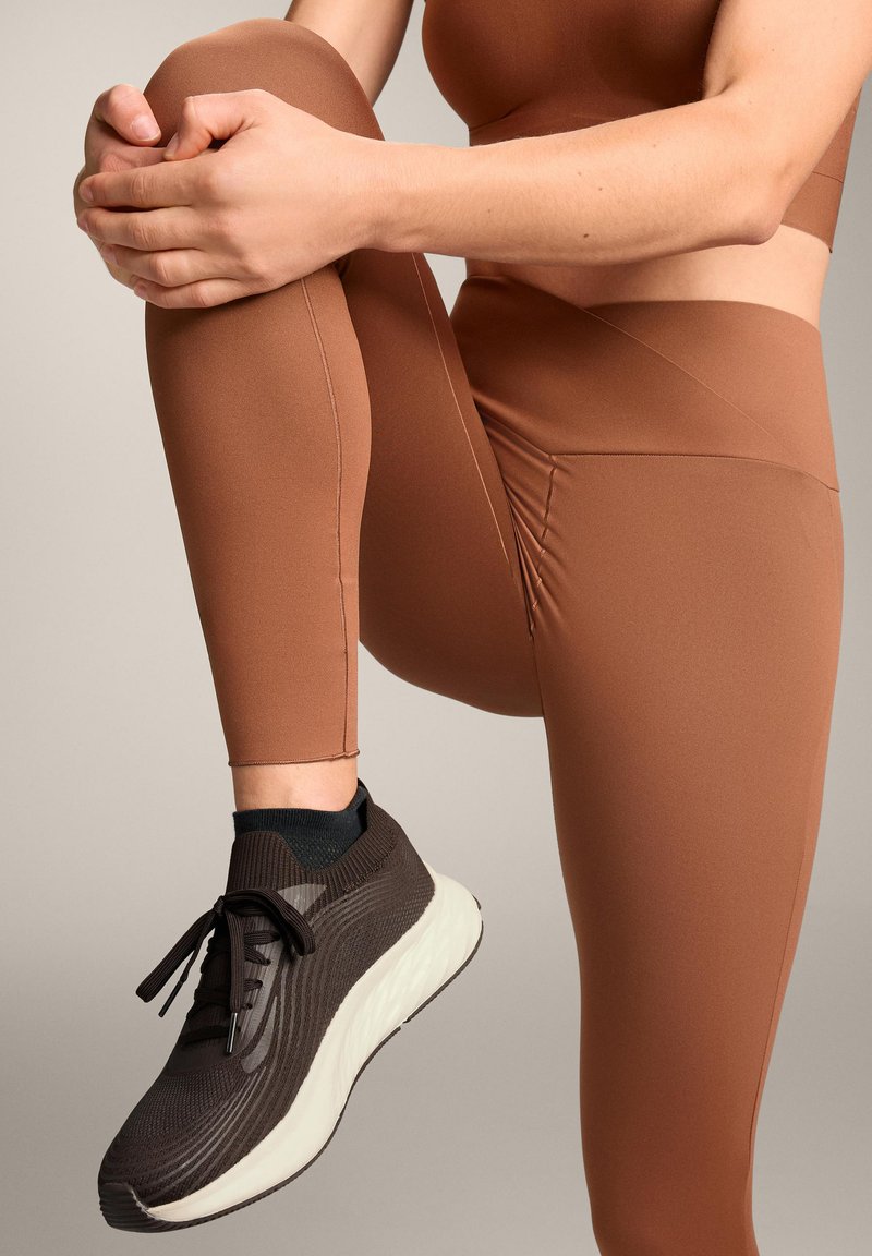 Bruine high-waisted leggings van glad materiaal, gecombineerd met een donkerbruine sneaker met een gestructureerde bovenkant en een witte zool.