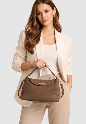 Femme portant un blazer beige et un pantalon, tenant un sac à main en daim marron avec une anse supérieure et une fermeture dorée, sur un fond uni.
