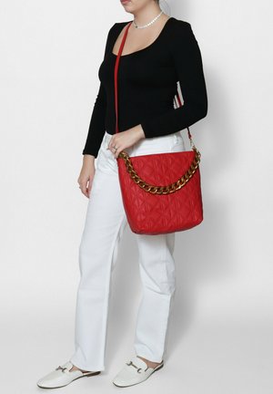 Borsa a tracolla rossa trapuntata con accentuazione in oro. Presenta una consistenza liscia e una tracolla, indossata con pantaloni bianchi e una maglietta nera.