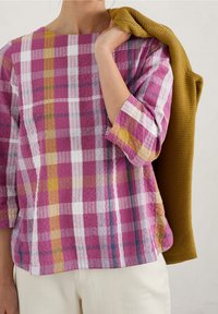 Purple and white checkered top with three-quarter sleeves, textured fabric, complemented by a mustard sweater draped over the arm.