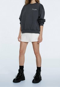 Sudadera de gran tamaño en gris oscuro con mangas enrolladas, combinada con una falda mini blanca y botas negras gruesas con cordones. Acabados en textil suaves visibles.