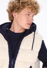 Fleecefodrad väst med en vit, texturerad utsida och en marinblå insida. Har en dragkedjestängning och huva, vilket ger en lager-på-lager-look.