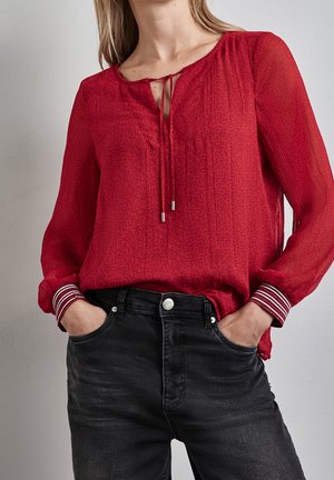 Vrouw draagt een rood blouse met lange mouwen en een strik bij de hals en zwarte spijkerbroek, handen in de voorzakken, tegen een effen achtergrond.