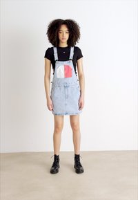 Vestido de tirantes de mezclilla con un lavado claro, que presenta un bolsillo delantero en rojo, blanco y azul. Combinado con una camiseta negra y botas negras.