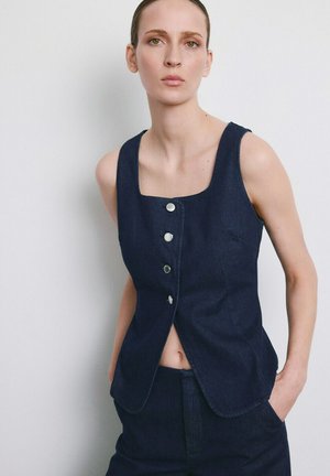 Mujer con blusa sin mangas azul oscuro abotonada y pantalones a juego, de pie con las manos en los bolsillos contra un fondo claro y liso.