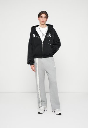 Zwarte rits hoodie met versleten letters, gedragen over een witte graphic t-shirt. Grijze sweatpants met een pluche zijstrip. Witte sneakers.