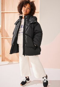 Schwarze Puffjacke mit hohem Kragen, glänzender Textur und Druckknöpfen, kombiniert mit einem hellgrauen Sweatshirt und weißen Hosen. Schwarze Sneakers.