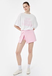 T-shirt bianco a maniche corte con lettering rosa, abbinato a una gonna rosa a portafoglio. La modella indossa sneaker bianche e calzini sportivi bianchi.
