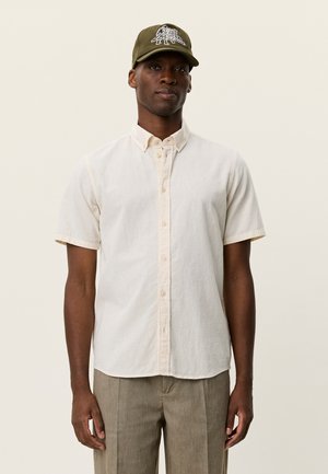 Kortærmet beige skjorte med knapper i tekstureret stof, kombineret med en grøn kasket med hvidt grafisk motiv. Modellen står med fronten mod kameraet.