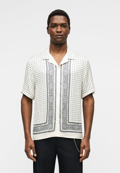 Άνδρας φοράει κοντομάνικο λευκό πουκάμισο με μικρά μοτίβα σε ναυτικό μπλε χρώμα αστέρια και παϊλέ, συνδυασμένο με μαύρο παντελόνι και αλυσίδα ως αξεσουάρ.