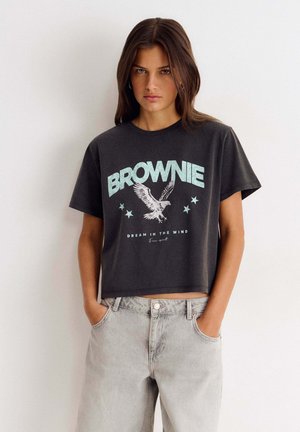 Jeune femme aux cheveux bruns raides porte un crop-top noir avec le texte "BROWNIE", un motif aigle et un jean taille haute gris clair, debout les mains dans les poches.