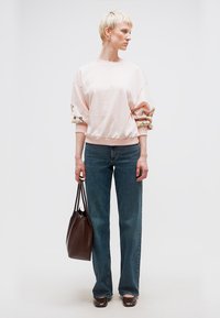 Γυναίκα με κοντά ξανθά μαλλιά που φοράει ανοιχτό ροζ φούτερ με φλοράλ μανίκια, μπλε τζιν, καφέ παπούτσια και κρατά μια σκούρα καφέ τσάντα ώμου.