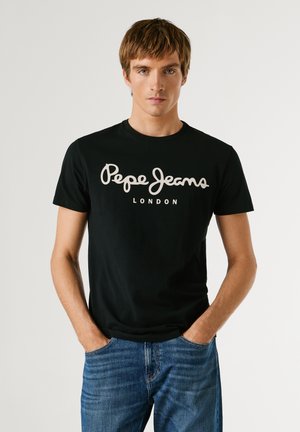 Jaunas vyras dėvi juodą Pepe Jeans London marškinėlį ir mėlynas džinsines kelnes, stovintis su rankomis kišenėse priešais paprastą foną.