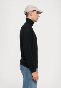 Svart polotröja med långa ärmar, tillverkad av slät tyg. Bärs med ljus denimjeans och en beige keps med justerbart bakband.