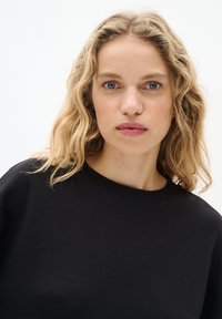 Felpa nera in cotone con collo rotondo. Texture morbida e vestibilità comoda. Indossata da una persona con capelli ondulati, lunghi fino alle spalle.