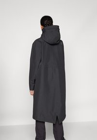 Langer schwarzer wasserdichter Mantel mit Kapuze, der eine taillierte Taille, elastische Bündchen und einen hohen Schlitz auf der Rückseite für mehr Bewegungsfreiheit bietet.