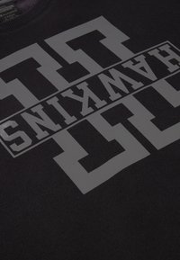 Sudadera negra con un gran estampado gráfico reflectante que presenta la palabra "HAWKINS" en letras mayúsculas en negrita, sobre un tejido texturizado.