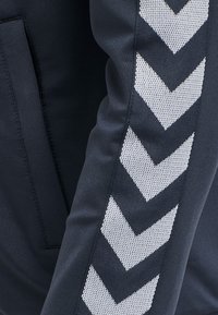 Marineblauwe sportieve trainingsbroek met een zijpaneel met wit chevronpatroon, gemaakt van textiel elastisch materiaal. Inclusief een zak.