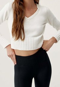 Kobieta z długimi, falującymi włosami nosi biały sweter na krótki rękaw oraz czarne legginsy z wysokim stanem, pozując z rękami na biodrach.