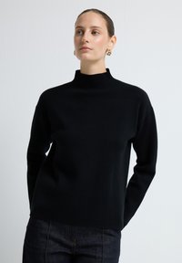 Kobieta w czarnym swetrze z długim rękawem i wysokim kołnierzem oraz ciemnych dżinsach, z złotymi kolczykami w kształcie kół, stojąca na tle jasnego tła.