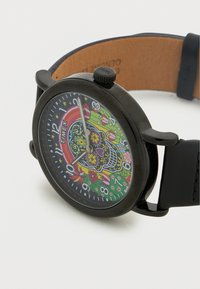 Montre Timex noire avec un cadran rond présentant un design de crâne coloré, des motifs floraux, et un bracelet en silicone noir. Les marqueurs numériques sont présents.