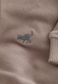 Tessuto di maglione grigio a maglia con una texture morbida. Presenta un piccolo logo di cammello ricamato grigio sulla parte anteriore, con dettagli a coste sui polsini.