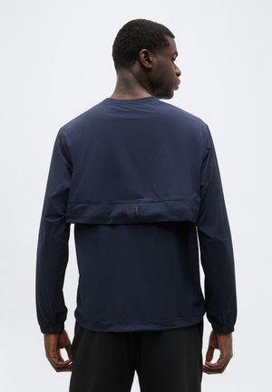 Homme de dos portant une veste bleu marine à manches longues avec un rabat aéré sur le haut du dos, se tenant devant un fond uni.