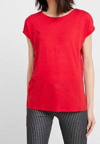 Mujer vestida con una camiseta roja de manga corta lisa y pantalones negros con rayas verticales blancas, de pie contra un fondo blanco.