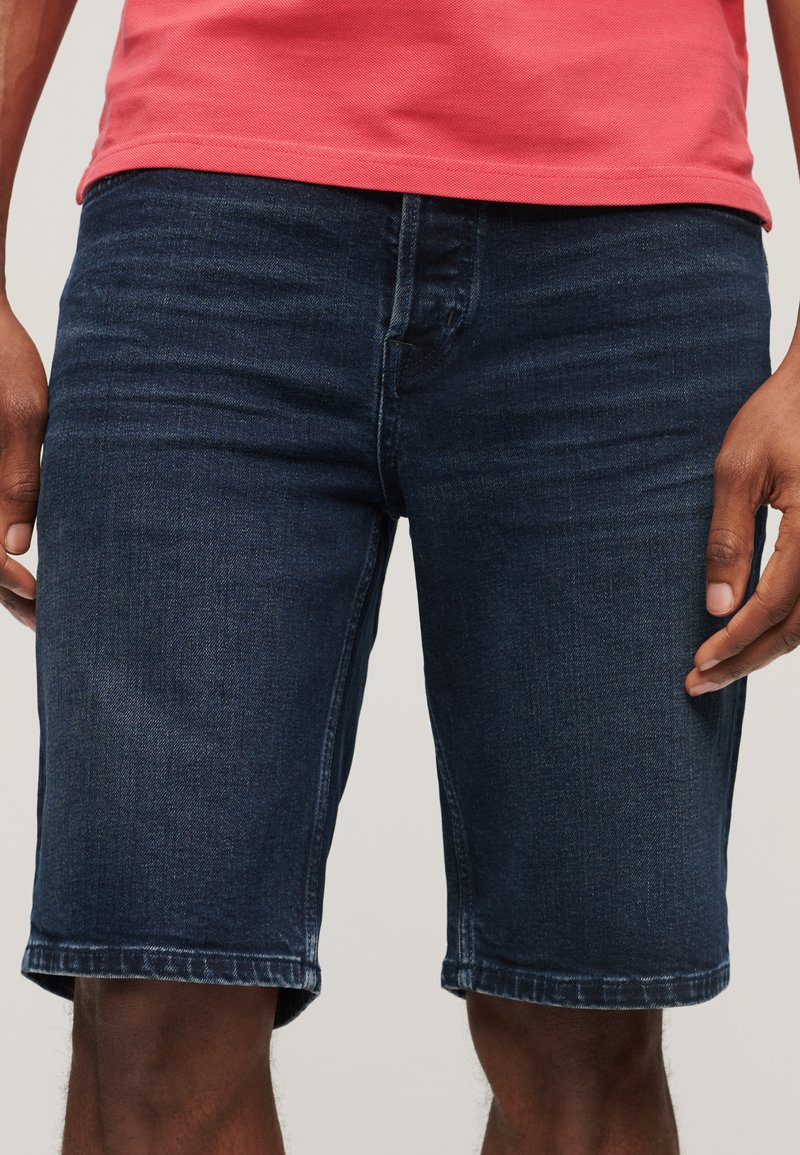 Granatowe szorty dżinsowe o prostym kroju, o długości średniej, z minimalnym wyblaknięciem. Posiadają pięć kieszeni oraz tradycyjne zapięcie na guzik.