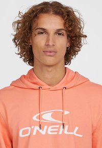 Korallefarbener Kapuzenpullover mit Kordelzügen und Kängurutasche. Auf der Brust befindet sich ein großes weißes Logo. Hergestellt aus weichem Stoff mit einem lässigen Schnitt.