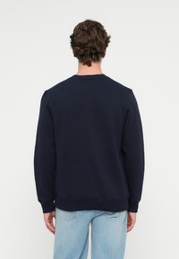 Námornícka modrá mikina vyrobená z bavlnenej zmesi, s okrúhlym výstrihom, rebrovanými manžetami a voľným strihom. Zobrazená zozadu s voľnými džínsami.