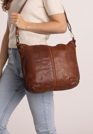 Femme tenant un sac à bandoulière en cuir marron, portant un pull crème à manches courtes et un jean bleu clair, sur un fond neutre.