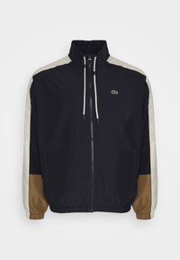 Marinblå jacka med vit och brun färgblockerade ärmar, hög krage, framdragkedja, elastiska manschetter och framträdande grön logotyp på bröstet.