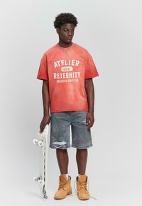 T-shirt rouge surdimensionné avec imprimé graphique blanc, associé à un short en denim déchiré et des bottes de travail beiges. Tient un skateboard blanc.