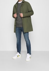 Olivově zelený kabát přes šedý svetr, doplněný modrými džínami a bílými teniskami se zelenými a žlutými pruhy. Hladká látka a střih na míru.